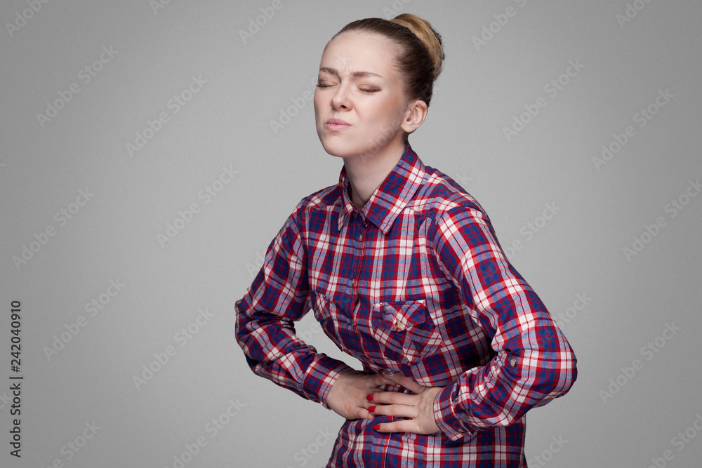 stomack sick unhappy girl in red, pink checkered shirt, collected bun hairstyle, makeup standing and touching her painful belly. indoor studio shot. isolated on gray background stomack sick unhappy girl in red, pink checkered shirt, collected bun hairstyle, makeup standing and touching her painful belly. indoor studio shot. isolated on gray background