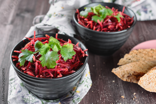 Thinly sliced ​​fresh beets in a black salad bowl with sprigs of cilantro ready for eating are on a wooden table. Shredded beet salad with toasts, closeup.