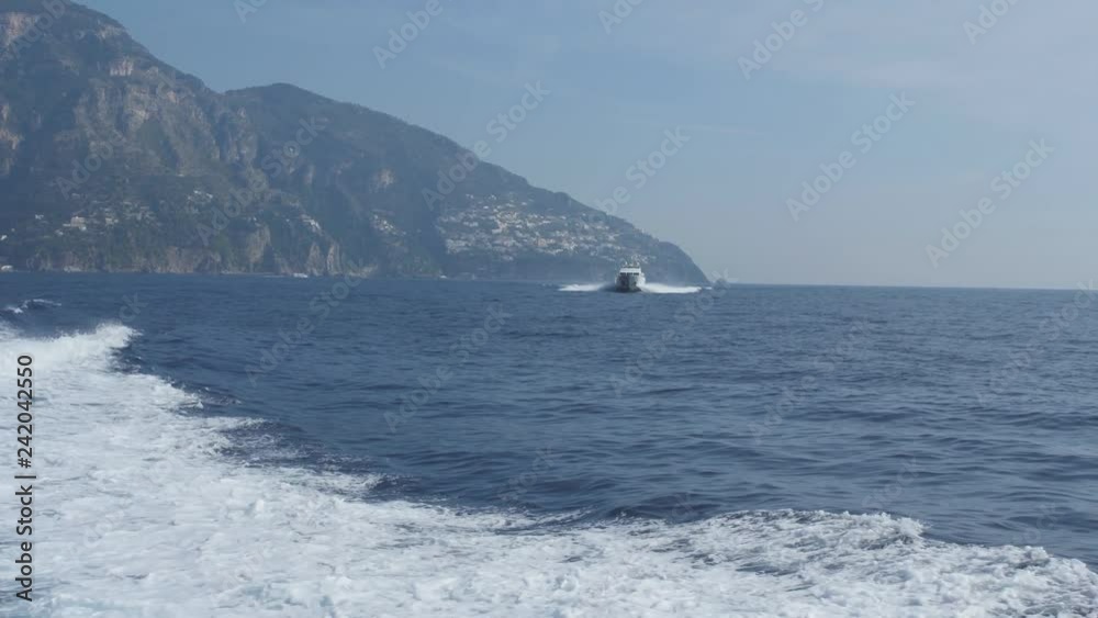 White foam track behind ship. Motor boat floats in blue sea against hilly shore. Positano, Italy White foam track behind ship. Motor boat floats in blue sea against hilly shore. Positano, Italy