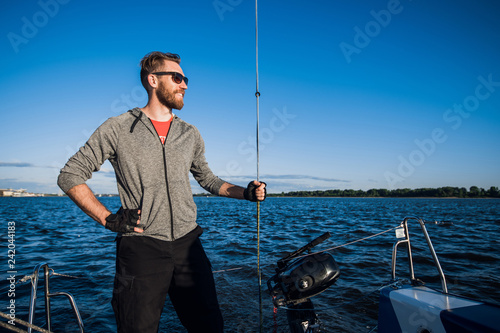 Young man on a catamaran standing and holding rope in front of a blue sky during cruising.