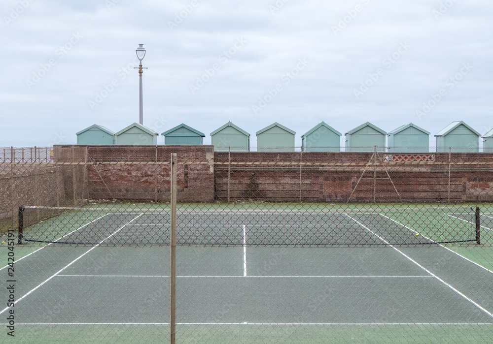 Tennis court, with row of green beach huts behind, on the sea front in Hove, Sussex, UK