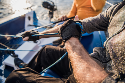 Close-up of male hands in gloves pulling rope of sailboat while mooring yacht and sitting with friends on deck.