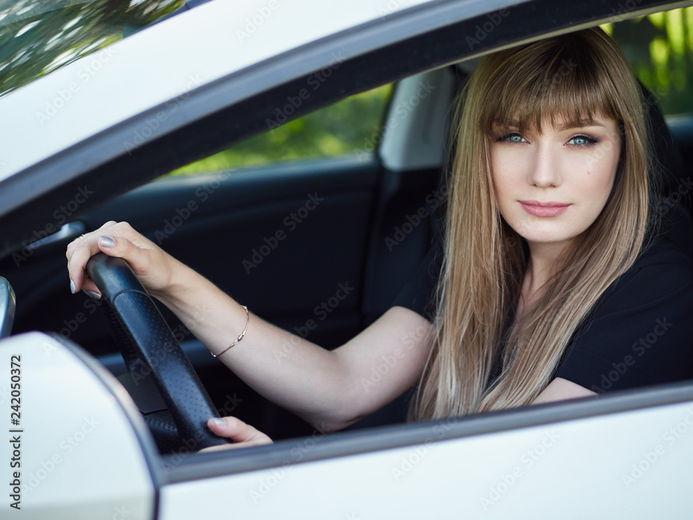 Happy young woman posing in her car holding steering wheel. Lady driver with long blond healthy hair at vehicle. Warm season lifestyle portrait.