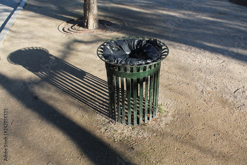 Metallic green trash bin, with metal grid screen, containing a black plastic garbage bag. Projecting elongated shadow. Located on the sand of the park. Image taken with daylight