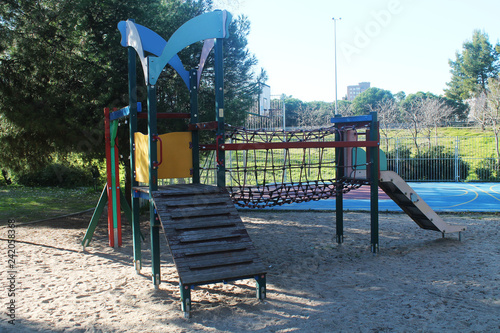 Playground for children, located in the park, consisting of a swing and suspension bridge. Colors red, green, blue and yellow. Located on sand. Image taken during day.