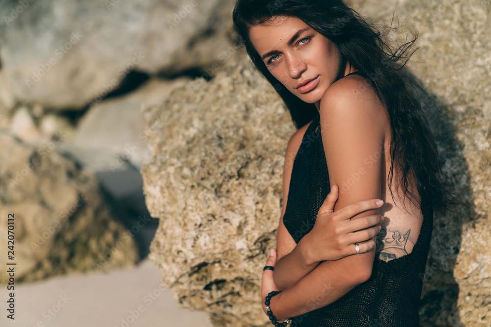 Portrait of sexy brunette model in knitted sweater posing on the sandy beach, big stones and rock on background.