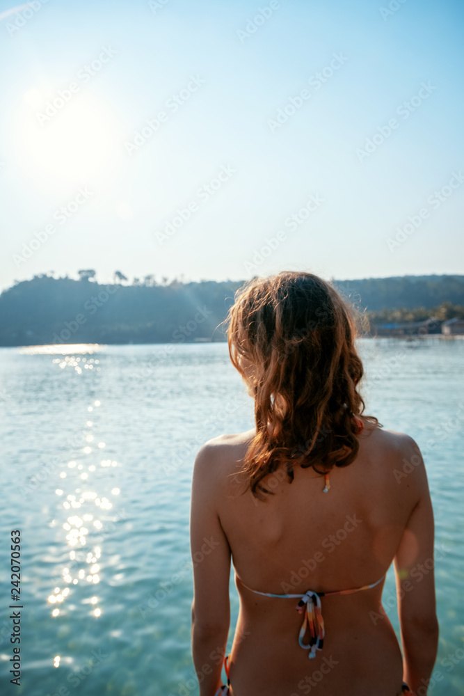 Young beautiful woman in bikini is standing back against the background of the sea, concept of leisure and travel to the tropics