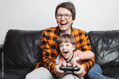 A little handsome boy and his dad are sitting on the couch at home and playing video games with the joystick. Dad and son have fun on a white background. They are happy and smiling