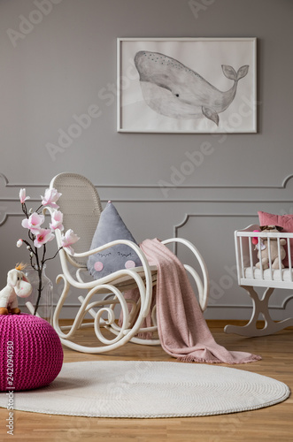 Pink pouf and flowers in grey baby's bedroom interior with poster and rocking chair. Real photo