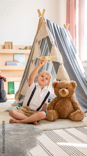 Cute little blonde boy sitting next to teddy bear in stylish scandinavian kid's playroom