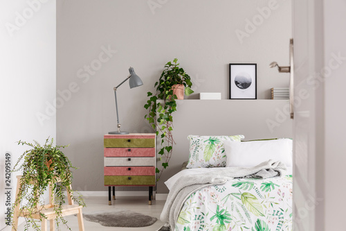 Urban jungle in grey scandinavian bedroom with wabi sabi nightstand and bed with floral duvet and blanket, real photo