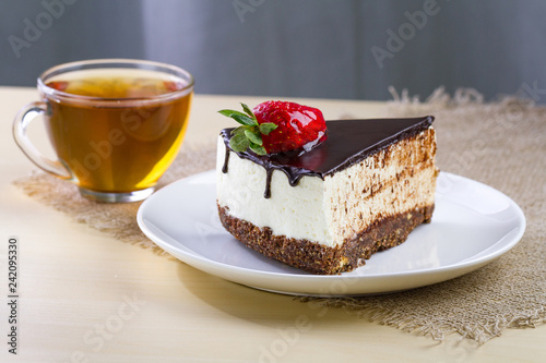A cup of hot tea and a slice of sweet cake with fresh strawberries and dripping chocolate glaze in a white plate. Sweet dessert