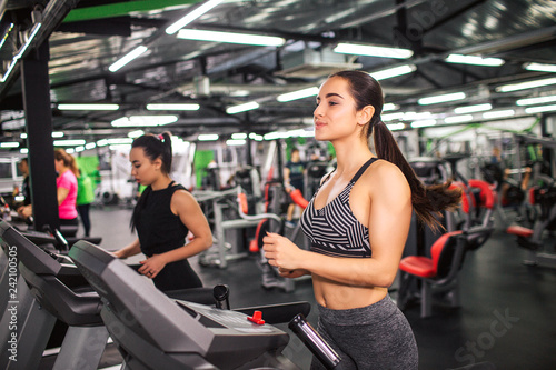 Nice young woman runs on running machine. She looks concentrated. Young asian woman runs as well further. They in gym room alone.