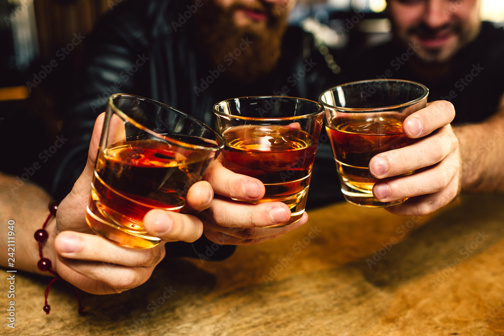 Cut view of three bearded young men holding glasses with rum together. They smile. People sit in bar. - obrazy, fototapety, plakaty 
