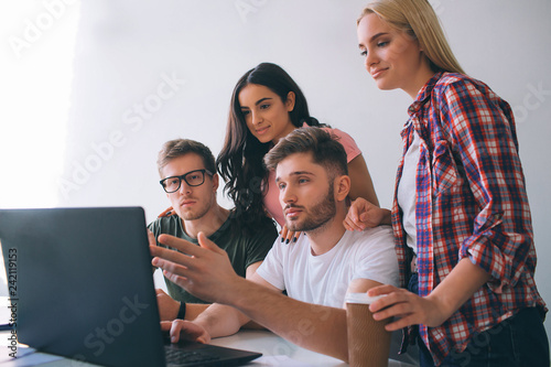 Serious young men sit at table and look at laptop screen. Brunette and blone young women stand behind them and smile. They are together in one room.