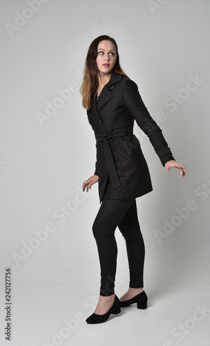 full length portrait of a brunette girl wearing long black coat, standing pose on grey studio background.