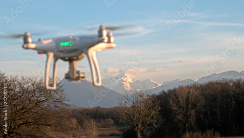 A drone hovers in flight in front of the mountains of the alps in France. The drone is stationary hovering above the ground as mountains appear in the distance.