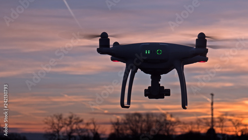 A drone hovers in flight in front of a sunset background. The drone is stationary hovering above the ground as the sunsets in the distance.