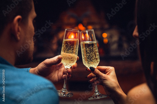 Young man and woman touchgin glasses with champaigne. They look at each other. Couple sit at fireplace. photo