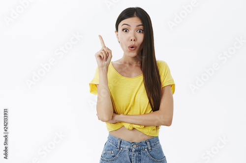 Amazed and excited attractive young slim female model in cropped yellow t-shirt with long dark hair folding lips raising index finger in eureka gesture  adding suggestion talking about her idea