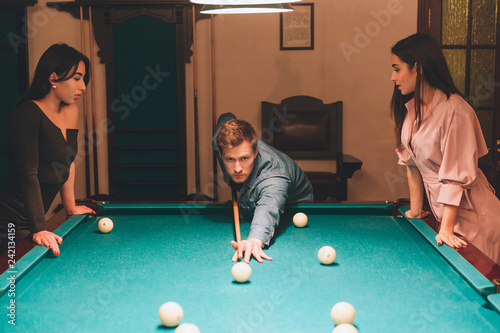 Young redhead player stand between two models. He aims into billiard ball. Young women stand and look at gamel. They lean to table. photo