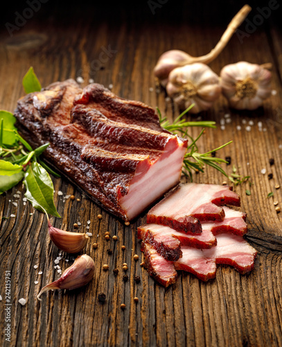 Smoked bacon, slices of smoked bacon on a wooden rustic table with addition of fresh aromatic herbs. Natural product from organic farm, produced by traditional methods