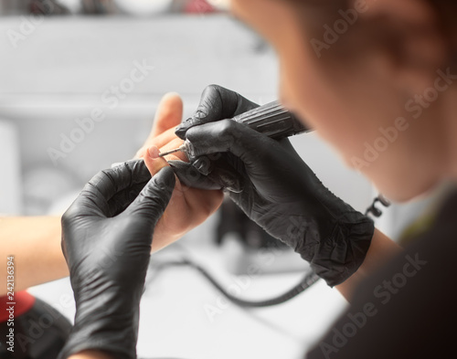 Process of manicure making in beauty salon. Close-up of professional master manicurist hands in black rubber gloves working on client fingernails with electric drill file. Fashion and beauty concept.