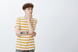 Portrait of calm and gloomy attractive young man with curly hair and tattoos leaning head on hand looking right with sad lonely look, being bored having nothing do standing upset and thoughtful