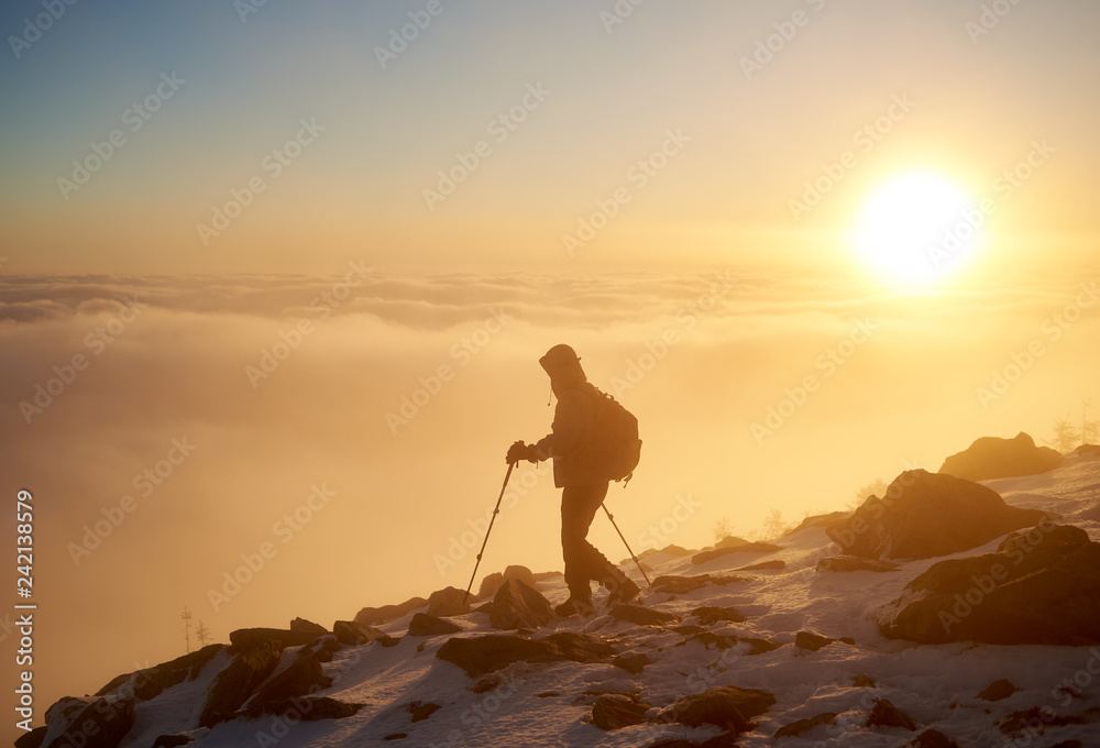 Tourist climber with backpack and trekking poles hiking on rocky snowy mountain steep slope on background of foggy valley filled with white puffy clouds, raising sun and blue sky at dawn.