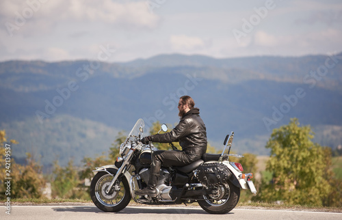Side view of bearded motorcyclist in sunglasses and black leather clothing riding cruiser motorbike along narrow asphalt path on sunny summer day