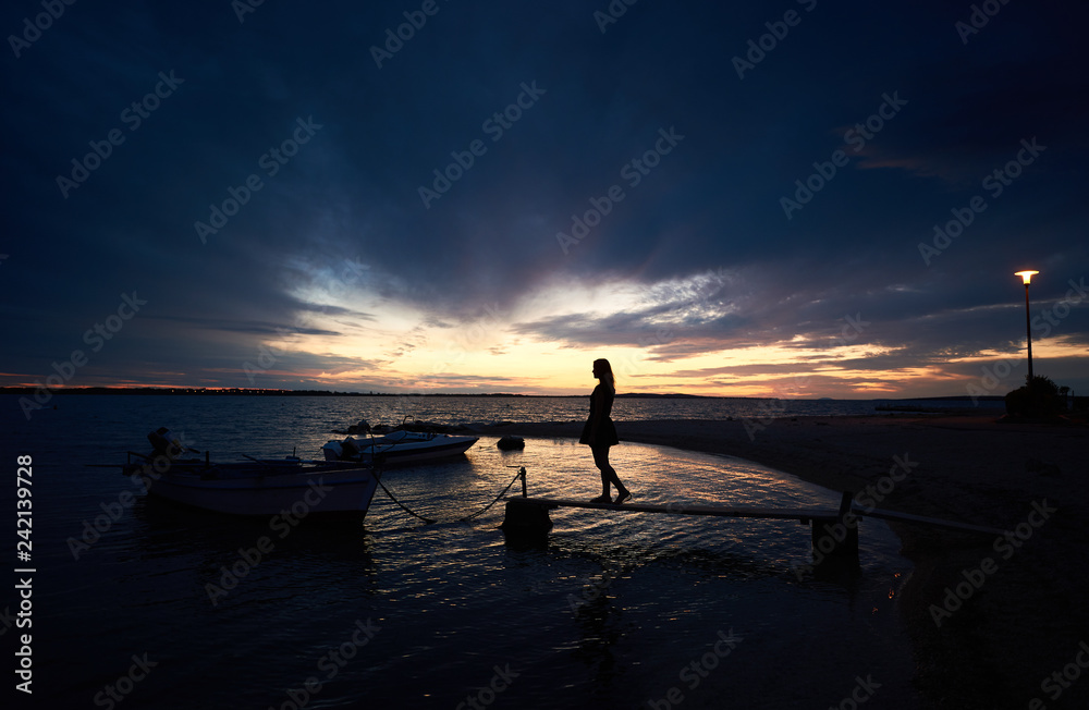 Fototapeta premium Silhouette of girl in short dress standing on narrow wooden bridge at small motor boats anchored at shore in sea inlet calm shallow water chained to pier on bright cloudy sky at sunset background.