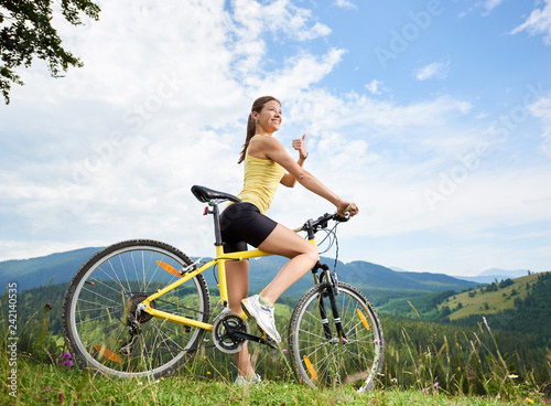 Attractive happy female cyclist riding on yellow mountain bike on a grassy hill, showing thumbs up, enjoying summer day in the mountains. Outdoor sport activity, lifestyle concept