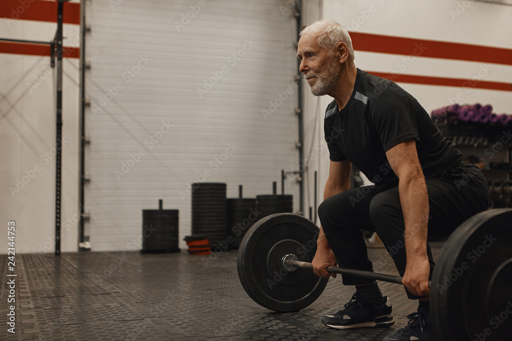 Stylish seventy year old male choosing healthy active lifestyle, training  with weights indoors, lifting barbell, bulding muscular strong arms, having  joufyl happy facial expression. Age and vitality Stock Photo
