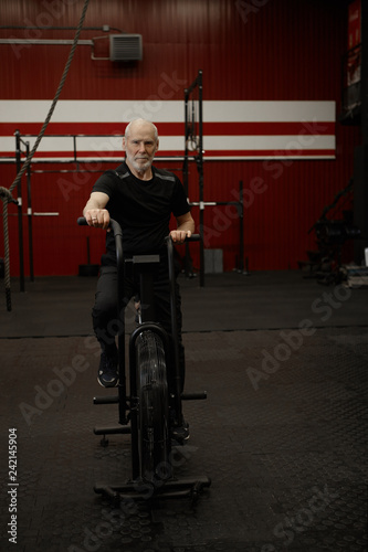 Vertical image of healthy self determined energetic elderly man with gray beard sitting on stationary bike, having cardio workout in gym. Senior male cycling on bicycle, wearing sports clohtes