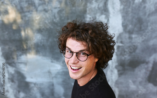 Top view portrait of handsome happy smiling male with curly hair, wears spectacles posing for social advertisement, isolated on gray concrete wall with copy space for your promotional information