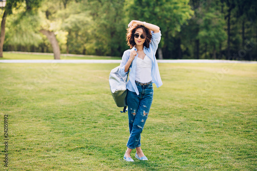 Full-length photo of cute brunette girl with short hair in sunglasses posing in park . She wears white T-shirt, blue shirt and jeans, shoes, bag. She is smiling to the camera.