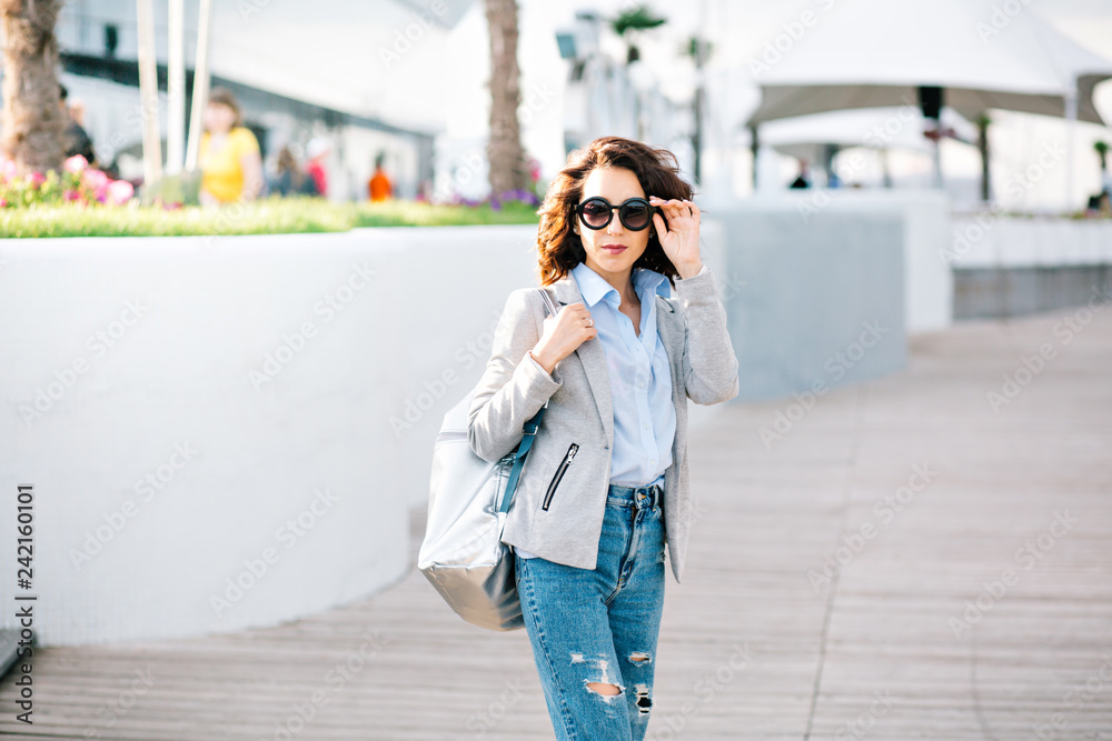 Pretty brunette girl with short hair is posing to the camera in city.  She wears shirt, jeans, jacket and bag. She holds sunglasses on face and looks to the camera.