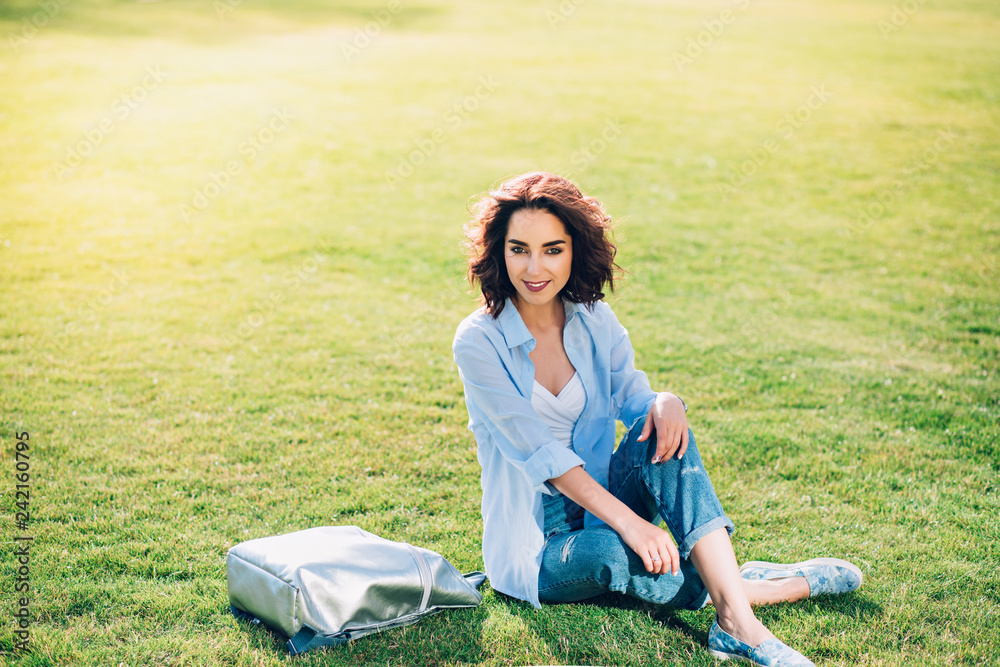 Nice brunette girl with short hair is posing on grass in park . She wears white T-shirt, shirt and jeans, shoes. View from above. Nice brunette girl with short hair is posing on grass in park . She wears white T-shirt, shirt and jeans, shoes. View from above.