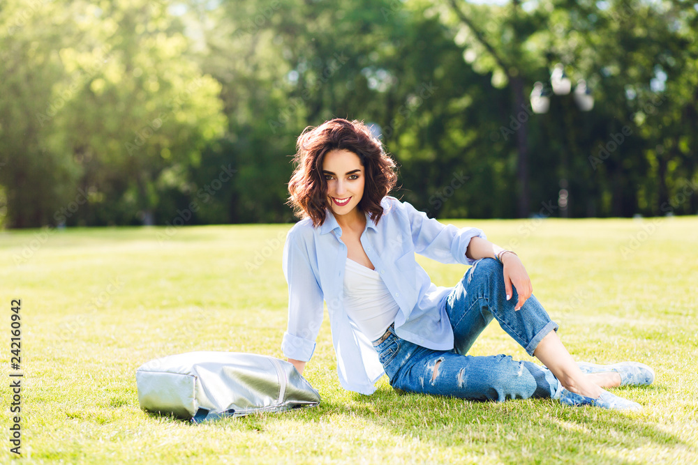 Full-length photo of cute brunette girl with short hair posing on grass in sunlight in park . She wears white T-shirt, shirt and jeans, shoes, bag. She is smiling to the camera. Full-length photo of cute brunette girl with short hair posing on grass in sunlight in park . She wears white T-shirt, shirt and jeans, shoes, bag. She is smiling to the camera.