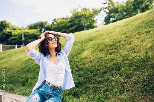 Attractive brunette girl with short hair in sunglasses is posing to camera in park on meadow background. She wears white T-shirt, blue shirt, jeans. She keeps hands on head and looks happy.