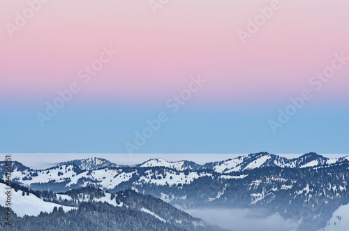 Forested hill country at a cold winter day in the Allgaeu Alps (Bavaria, Germany). Dawn with blue and red illuminated sky. Copy space.