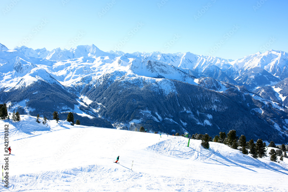 People enjoying skiing on prepared slopes in the Alps on sunny day. Beautiful snowy trees in the mountains. Perfect winter holidays destination for family in modern comfortable Alpine ski resort. 