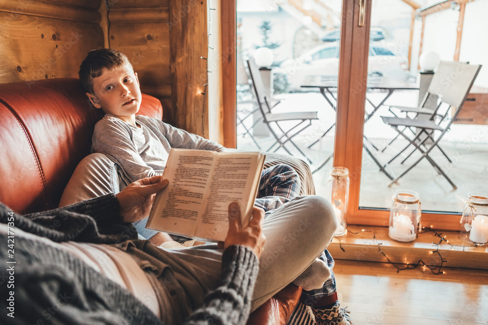 Father and son reading book together lying on the cozy sofa in warm country house. Reading to kids conceptual image.