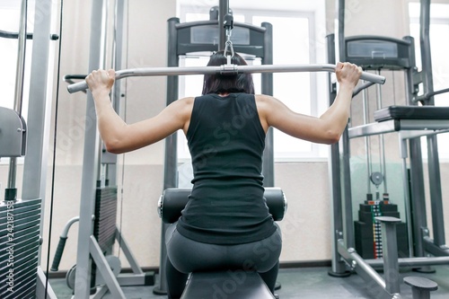Young woman doing exercises for the back on a fitness machine in the gym, view from the back. Fitness, sport, training, people concept.