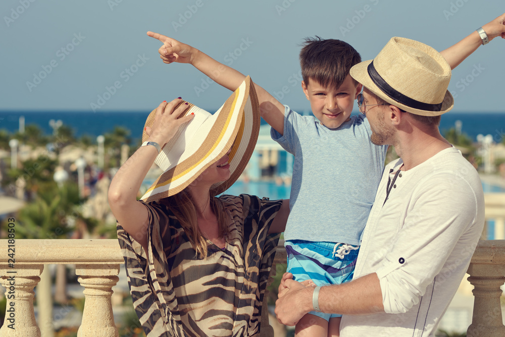 Family of three people on hotel balcony against sea in summer enjoying their vacation. Fathers is holding son on hands.
