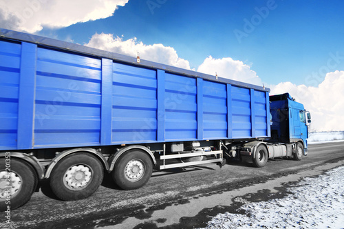 Truck with a trailer on the winter countryside road with snow against blue sky