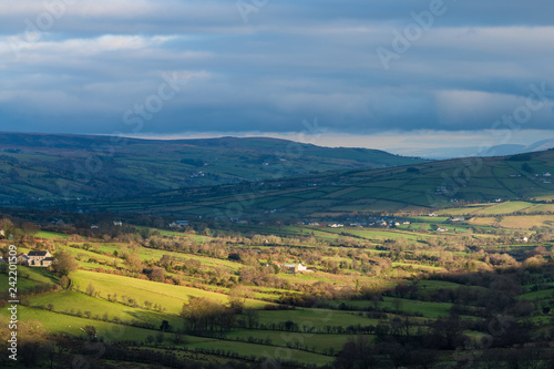 Bright sunlight breaking through clouds highlights a beautiful lush green landscape of farms and fields in Glenariff, County Antrim, Northern Ireland