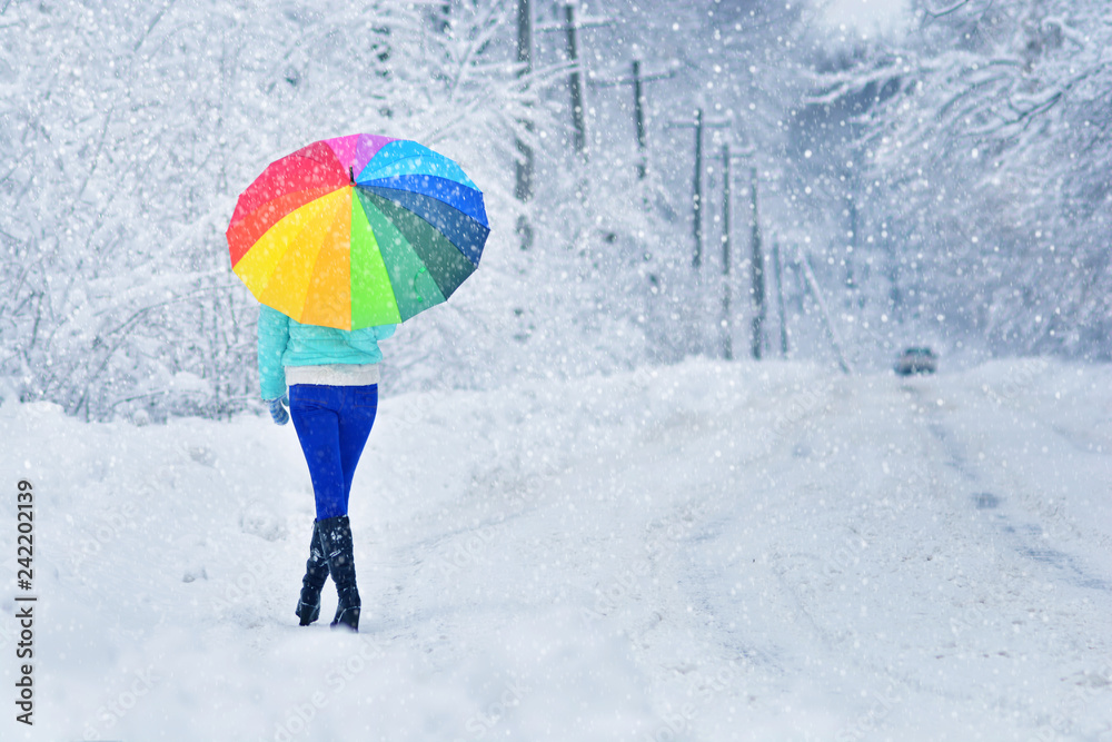 Woman with a rainbow umbrella on country road waits a car. Winter background. Positive concept.