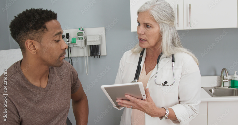 Fototapeta premium Mature white female doctor using portable computer pad to take patient notes. Young male african-american patient meeting with senior doctor indoors health clinic