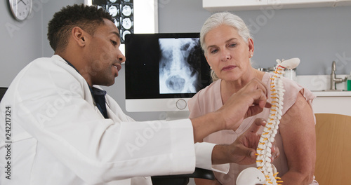 Senior female patient consulting with young doctor about back injury. Close up of black doctor holding model of spinal cord and explaining to elderly woman source of her pain